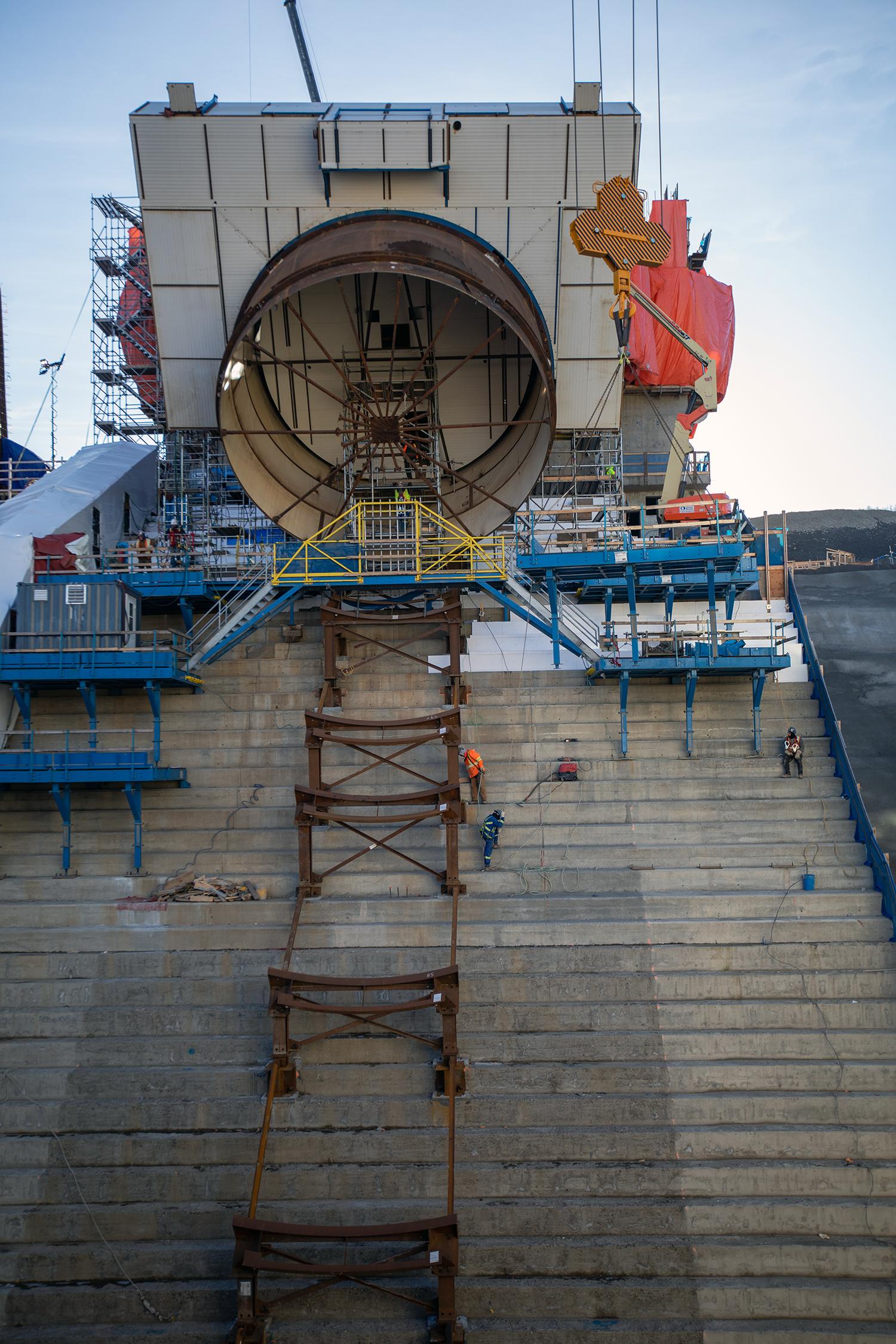 The square, white collar around the penstock is an external scaffolding to protect welders while they join the seams. More than 40% of the welding is complete and 13 of 18 pieces needed for this penstock are on site and in position. | October 2019