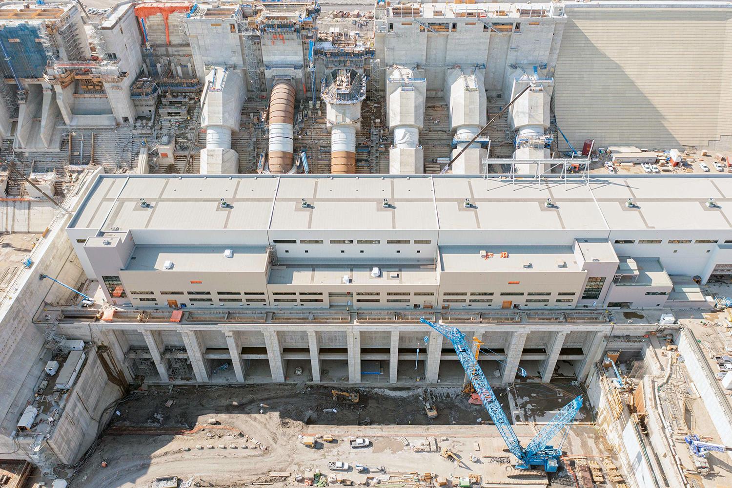 From left, penstock units six through one behind the powerhouse and operations building. Foundation enhancement work is ongoing at the tailrace in front. | August 2022