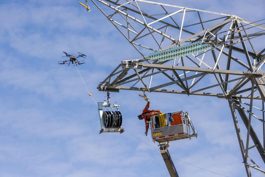 The drone pulls the pulling line while a powerline technician stands by to help guide the line through a sheave attached to the tower. | May 2024
