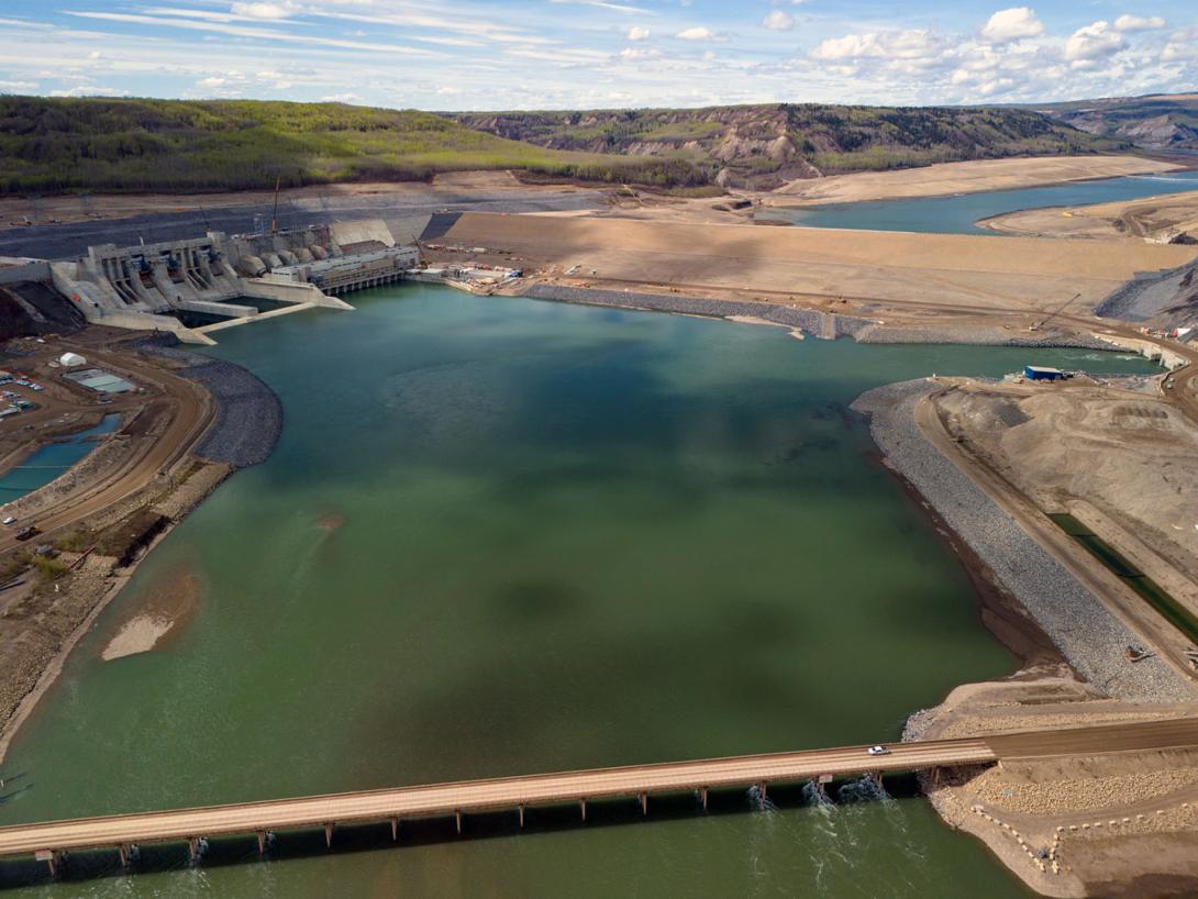 An upstream view of the completed Site C dam and generating station. The Peace River exits the diversion outlet portal at top right. | May 2024